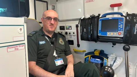 Paramedic Garry Cornelius-Dodds sitting in a chair in the back of an ambulance. He is in a dark green uniform and he is smiling at the camera. He has a bald head and is wearing glasses.