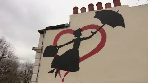 The mural shows Mary Poppins holding an umbrella in her left hand and a carpet bag in her right, flying towards a real-life chimney in the outline of a red heart.