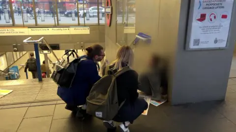 A male with a ponytail and classes and a female with shoulder-length blond hair crouch down to speak to a person sitting on some newspaper at the top of some stairs.