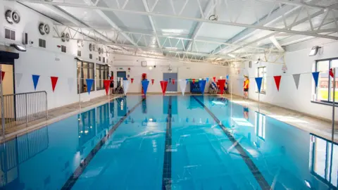 A photo taken at the foot of an indoor swimming pool. It has four lanes. Above the pool, from left to right, are two pieces of bunting.