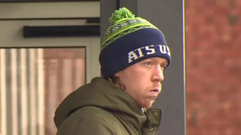 William Brown. He is wearing a green and navy bobble hat and a khaki green coat. 