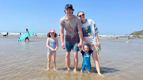 Family handout The image shows a family of four standing on the shore on a beach. It is a summer's day and the two children - aged one and five - are in their swimming costumes. The husband and wife are wearing shorts and t-shirts. They are all looking at the camera and smiling. 