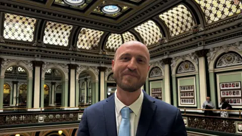 A man in a suit stands inside a council chamber