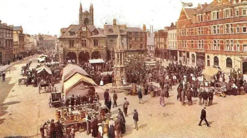A picture of the market in Peterborough's Cathedral Square with a monument in the middle and crowds of people around covered market stalls.