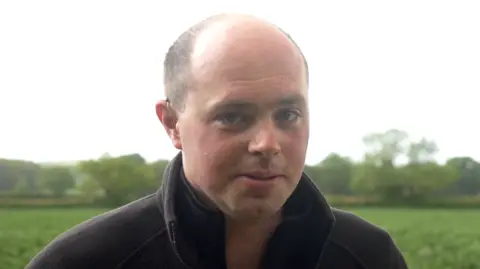 Matthew Register has a shaved head and is wearing a black jumper in front of a green field.