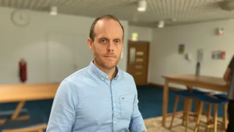 A man in a blue shirt sits at a desk in an office, there are tables and stools in the background