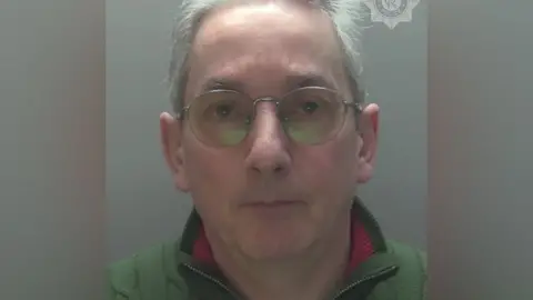 Police custody photo of Andrew Pennington, who has receding grey hair and blue eyes. He is wearing metal-framed specs on his face, a green fleece with a red trim on the collar.