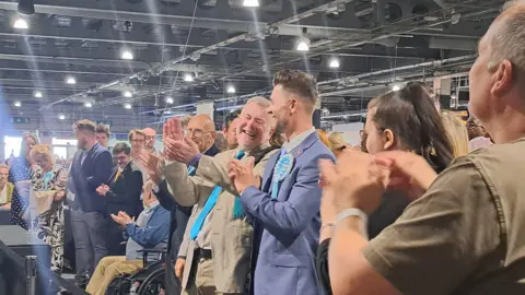 A crowd of Reform UK supporters, many wearing turquoise ties and rosettes, are cheering and clapping at an election count