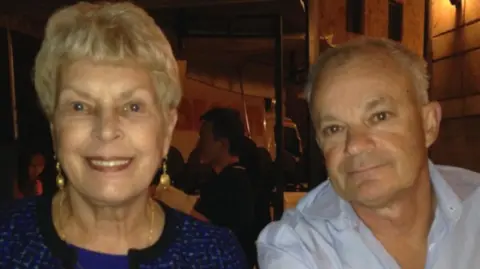 Simon Rendell A woman and man sit together in a restaurant. The woman has short light coloured hair and wears a blue cardigan, top and pearl earrings that hang down. The man has short, thin grey hair and wears a blue shirt.