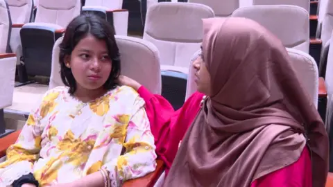 Nure Jannat Yusha, with short black hair and wearing in a floral dress, sits beside a woman in a pink hijab. 
