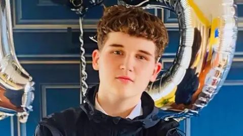 Head and shoulders shot of a teenage boy with brown wavy hair, combed forward, and brown eyes. He is staring directly at the camera with his head tilted slightly the left. He is wearing a dark blue jacket, zipped up to the collar, and a white shirt underneath. The boy is standing against a blue wall and two silver helium balloons - a 1 and a 5 - are partially visible in the background. 