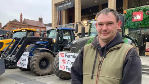 Will Morgan has short brown hair. He is wearing a green gilet and a black hoodie. He is standing in front of several tractors which have signs on them reading "no farming, no food, no future".