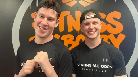 Shay and Bobby are both wearing black T-shirts and caps facing backwards. They are standing in front of a black and orange Fitness By Bobby logo.