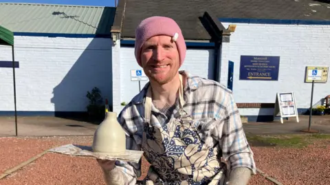 A man with a pink beanie on and a ginger beard. He is wearing a beige and blue check shirt with a similar coloured floral pattern apron over the top. He is holding a miniature bottle kiln which he has just finished throwing.