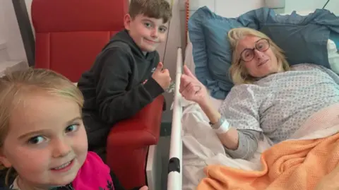 Helen in a hospital bed with her two grandchildren standing next to her. She wears a hospital gown and has a orange blanket over her. A young boy sits on a red chair next to her with his thumb up, and a young girl with blonde hair smiles at the camera.