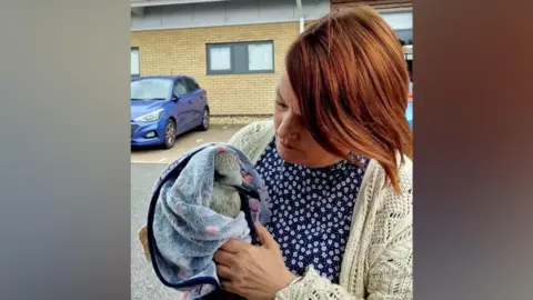 Supplied A woman wearing a cream coloured cardigan over a flowery top holding a seagull wrapped in a towel. She is looking down towards it.