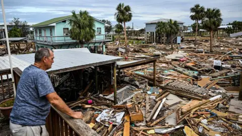 CHANDAN KHANNA/AFP Seorang pria memeriksa kerusakan rumahnya pasca Badai Helene. Gambar tersebut menunjukkan puing-puing dari rumah-rumah sejauh mata memandang.