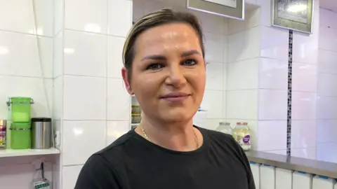 Dorina Gjoshi standing behind a fish shop counter against a white tiled wall background wearing a black top and hair tied back. 