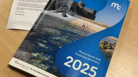 A 2025 phonebook with a blue cover and a picture of a castle and a sandy beach on it. It also features the Manx Telecom logo and the words Phone book and business directory 2025 on the front.