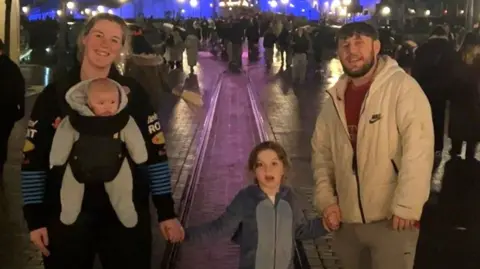 Ruairi George O'Connor A woman, with a baby strapped to her front, holding hands with her five-year-old daughter. The girl is also holding hands with her father, who is wearing a red top and white coat. He has short brown hair and a brown beard. 