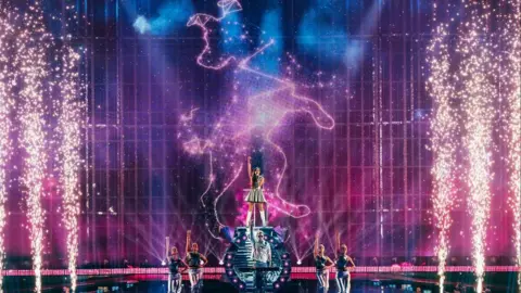 Sarah Louise Bennett / EBU A stage setting with a performer in a silver dress raised on a podium, there are lots of pyrotechnic sparks either side, with two dancers on either side.
The outline of a dog, styled like a star constellation, is projected on the wall behind with purple lighting