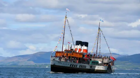 Sylvia Watson The boat 'Waverley' sailing through water, with its famous black, white and red funnels, and bunting and flags, with hills in the distance, under a cloudy blue sky.