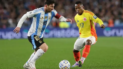 Getty Images Lionel Messi, wearing the blue and white striped shirt, black shorts and white socks of Argentina, prepares to dribble past an opponent who is wearing the yellow shirt, white shorts and red socks of Colombia.