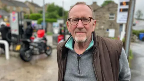 BBC/Seb Cheer A man, David, smiles at the camera. He is wearing glasses, a grey jumper and a brown gilet. He is standing next to a petrol station forecourt, where motorcyclists are refuelling.