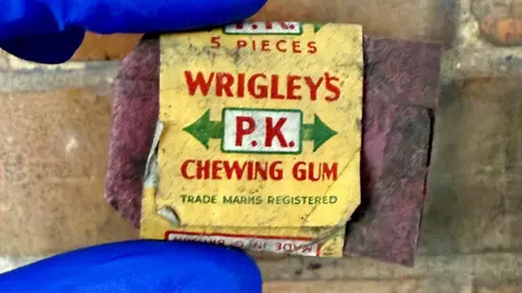 North Northamptonshire Council An old and empty Wrigley's yellow chewing gum wrapper with red writing and green arrow markings, is being held up by a person wearing blue gloves by a brick wall. 