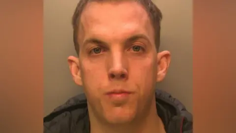 A mug shot of a man with short, brown hair. He is wearing a black coat.
