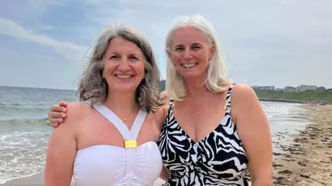Two grey-haired women in swimsuits smile at the camera, a view of the beach behind them. The woman on the left has shoulder length wavy hair, and wears a white swimming costume with gold jewellery. The woman on the right has shoulder-length straight hair and wears a black and white patterned costume.