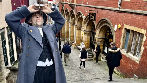 A performer from the Invisible Circus, dressed as an air raid warden, uses old fashioned binoculars to pretend to look for German aircraft in the sky. He is wearing a long grey coat and a protective helmet