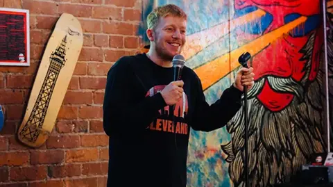 Kieran Cooper A man wearing a black jumper with short light brown hair is holding a microphone and smiling. He is on a stage with some wall decorations behind him including a wooden image of the Blackpool Tower.