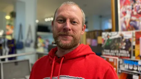 BBC Lee Evans has short brown hair and is wearing a red hoodie. He is standing inside the vintage shop filled with clothes, bags and posters