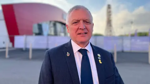 William Irvine looking at the camera. He is wearing a navy suit and tie, and a white shirt.