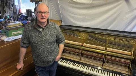 The picture shows a middle-aged white man standing inside a busy workshop filled with tools, boxes and piano parts. The man is leaning against an upright piano with a wooden exterior on the left side of the image. Next to them is another upright piano with its front panel removed, revealing the metal frame, strings, hammers and action mechanism. 