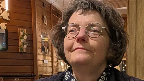 This picture shows a person seated at a table inside what looks like a cosy, warmly lit restaurant or café with a strong wooden interior. The space has wood-panelled walls, wooden ceiling slats and soft hanging lights that give the scene a relaxed, intimate feel. Decorative details are visible in the background, including framed items on the walls and small ornaments hanging from the ceiling.