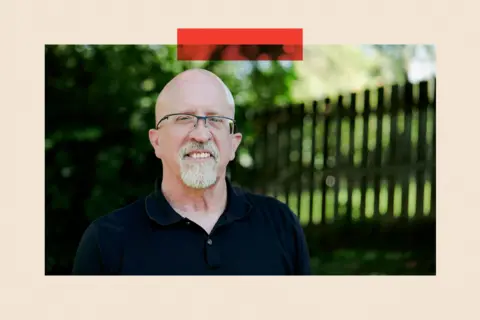 A white man with a bald head, beard and glasses stands smiling in front of a black fence with green trees behind it