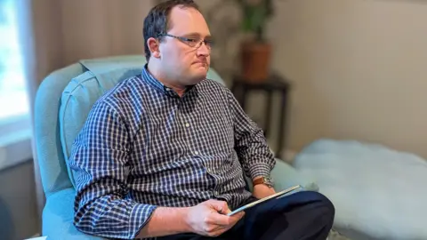 Nicholas Strahl, wearing glasses and a checked shirt, sits in an armchair holding a tablet.