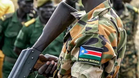 A close up of a soldier's arm showing a South Sudan flag on his shoulder - his face is not shown. The butt of his rifle is also visible.