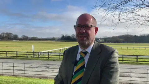 BBC/Nathan Turvey The CEO of the Yorkshire Agricultural Society, Allister Nixon, wearing glasses, a shirt and tie and suit jacket, with fields and fencing behind him on a sunny day.