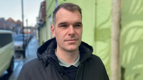 Michael Harnett, stepfather of Christie Harnett. He is standing on a pavement, next to a green-painted building. He has short brown and grey hair and blue eyes. He is wearing a green polo shirt under a navy jumper and dark coat. He is looking in the distance. The background is blurred.