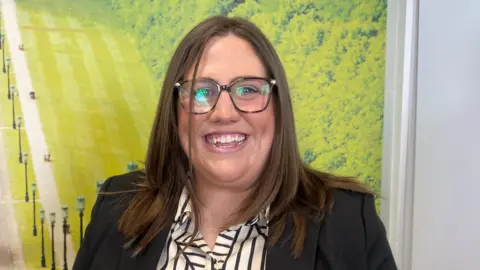 Julie smiling at the camera. She has long brown hair and is wearing glasses, a white and black striped shirt and a black blazer.