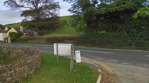 Google The junction at the White Hoe pumping station, showing the road running past it. There are green field and bushes across the road and a stone wall to the left next to a grass bank.