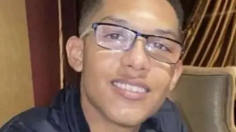 Family handout A man with short black hair, wearing glasses and a black jacket. He is sitting in a beige-coloured chair.