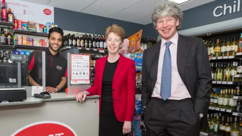 PA Than Thevarajah stands behind the till in his post office to the right of smiling Paula Vennells and Tim Parker with shelves stocked with alcohol in the background