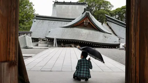 A woman with an umbrella squats in front of a collapsed shrine on Wednesday in Yatsushiro