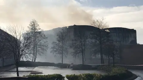 A large building destroyed by fire with smoke surrounding the premises. There are trees in front and police tape around the building.