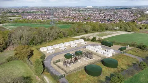 A mocked-up image of how the battery storage facility could look if plans get the go-ahead. It is an image of farmland with a residential area behind it. In the middle of the farmland a series of white rectangular buildings alongside large green storage structures, some circular, some rectangular, across the same site. 
