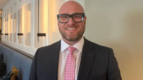 A man in a black suit with white shirt and pink tie. He is bald and wearing black glasses.